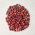 A pile of speckled Whipple bush bean seeds in shades of tan, brown, and maroon on a textured white surface.