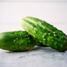 Two fresh, bumpy green Bushy cucumbers resting on a textured gray surface against a white background.