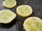 Thinly sliced yellow Lemon cucumber rounds with visible seeds resting on a dark, textured surface.
