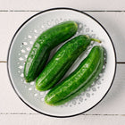 Three fresh, green Muncher cucumbers resting in a white decorative colander on a white wooden surface.