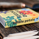 Edible Petals seed kit in a yellow matchbox-style package with white birch garden markers on a dark wooden surface.