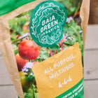 Gaia Green All-Purpose 4-4-4 fertilizer bag with green branding and a garden scene featuring tomatoes on a wooden surface.