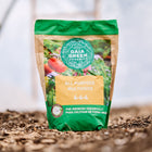 Gaia Green All-Purpose 4-4-4 fertilizer bag standing upright on soil in a greenhouse setting.