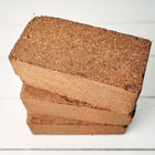 Three compressed brown Coco Coir Bricks stacked on a white wooden surface.