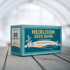 Teal cardboard box labeled 65-packet Emergency Heirloom Seed Bank sitting on a wooden surface in a greenhouse.