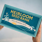 Hands holding a blue rectangular Emergency Heirloom Seed Bank box with gold text and a farm landscape illustration.