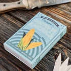 Rooted in America seed collection box featuring corn illustration on a rustic wooden table with garden markers.