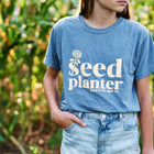 Blue garment-dyed Seed Planter short sleeve t-shirt with a white graphic, worn with denim shorts in a garden.