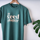 Forest green Seed Planter short sleeve cotton t-shirt with cream-colored graphic text on a wooden hanger.
