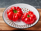 Two ripe, red, tomato-shaped Sheepnose Pimento peppers resting in a white enameled colander on a wooden board.