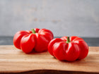 Two ripe, red, tomato-shaped Sheepnose Pimento peppers resting on a wooden cutting board.