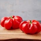 Two ripe, red, tomato-shaped Sheepnose Pimento peppers resting on a wooden cutting board.