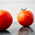 Two bright red, dew-covered Bonny Best tomatoes resting on a dark, reflective surface against a white background.