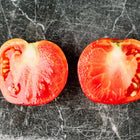 Two halves of a bright red Bonny Best tomato showing juicy, seeded flesh on a dark, scratched stone surface.