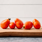 Five red, plum-shaped Italian Roma tomatoes lined up on a wooden cutting board against a white shiplap background.