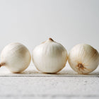 Three globe-shaped White Sweet Spanish onions with papery white skin on a white wooden surface.