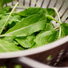 Fresh green Roquette arugula leaves in a stainless steel colander on a wooden surface.