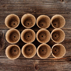 Twelve 3-inch round CowPots made of brown, fibrous composted material arranged on a dark rustic wooden surface.