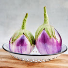 Two round, lavender and white Rosa Bianca eggplants with green stems sitting in a white colander on a wooden surface.