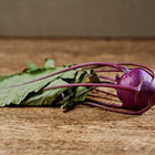 A single harvested Purple Vienna kohlrabi bulb with green leaves resting on a rustic wooden surface.