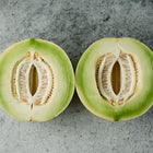 Two halves of a Tam Dew honeydew melon showing pale green flesh and seeds on a grey stone background.
