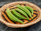 Fresh green Clemson Spineless okra pods arranged in a round woven bamboo basket on a dark marble surface.