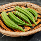Fresh green Clemson Spineless okra pods arranged in a round woven bamboo basket on a dark marble surface.