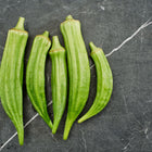 Five fresh, green Clemson Spineless okra pods arranged vertically on a dark grey marble surface.
