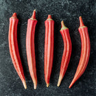 Five slender Jing Orange okra pods with vibrant red-orange skin arranged on a dark, textured background.