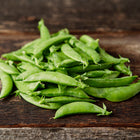 A pile of fresh, bright green Sugar Daddy snap pea pods resting on a rustic dark wooden surface.