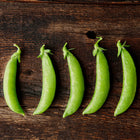 Five fresh, glossy green Sugar Daddy snap pea pods lined up on a rustic dark wooden surface.