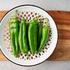 Fresh green Anaheim peppers in a white decorative colander on a wooden cutting board.