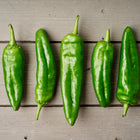 Five long, smooth green Anaheim peppers arranged in a row on a grey wooden surface.