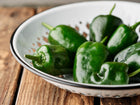 Fresh dark green Poblano peppers in a white enamel colander on a rustic wooden surface.