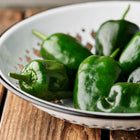 Fresh dark green Poblano peppers in a white enamel colander on a rustic wooden surface.