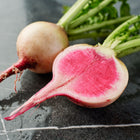 Whole and halved Watermelon radish showing pale skin and a vibrant rose-pink interior on a dark stone surface.