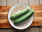Two Cocozelle zucchini with light green stripes resting on a white decorative colander atop a wooden board.