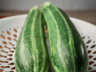 Two Cocozelle zucchini with light and dark green striped skin resting in a white decorative bowl.