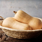 Two smooth, tan Waltham Butternut squash resting in a woven straw basket on a rustic wooden table.