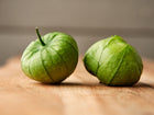 Two fresh Verde tomatillos with papery green husks resting on a wooden surface.