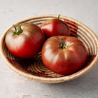 Three mahogany-red Black Krim tomatoes with green shoulders resting in a woven decorative bowl.