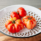 Three ribbed, rosy-red Rosa Sicilian tomatoes resting in a white decorative metal bowl on a wooden board.