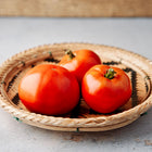 Three ripe, round Moskvich red tomatoes resting in a shallow, woven wicker basket on a gray surface.