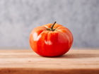 A single, round, red Brandywine tomato with a small stem resting on a wooden surface against a gray background.