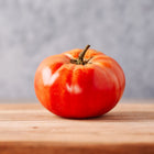 A single, round, red Brandywine tomato with a small stem resting on a wooden surface against a gray background.