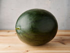 Round, dark green Sugar Baby watermelon sitting on a light wooden surface against a grey background.