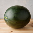 Round, dark green Sugar Baby watermelon sitting on a light wooden surface against a grey background.