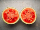 Two halves of a Sugar Baby watermelon showing bright red flesh and dark seeds on a grey textured surface.