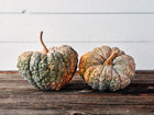 Two ribbed, warty Chirimen winter squash with green and buff skin sitting on a rustic wooden surface.